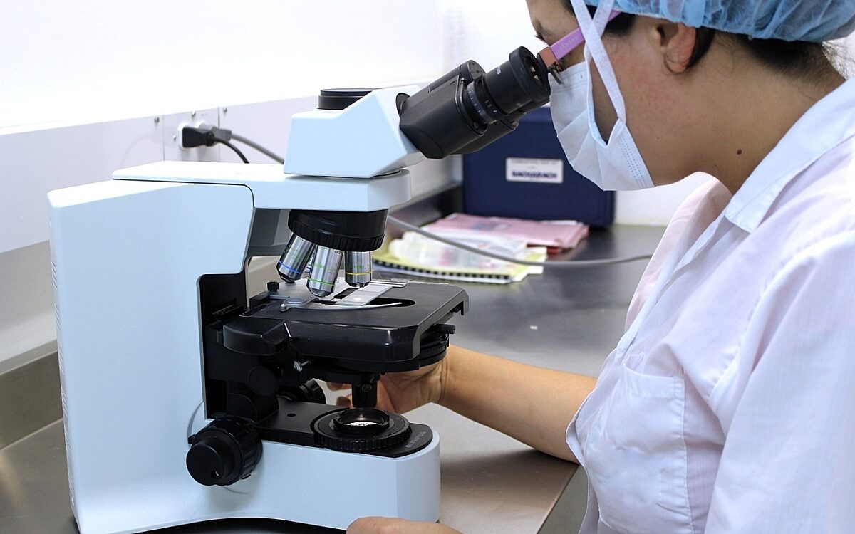 Eine Frau in weißem Kittel und mit Haar- und Mundschutz blickt in einem Labor in ein Mikroskop Eine Frau in weißem Kittel und mit Haar- und Mundschutz blickt in einem Labor in ein Mikroskop