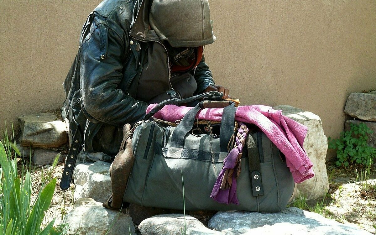 Eine obdachlose Person sitzt mit einer Reisetasche vor einer Mauer Eine obdachlose Person sitzt mit einer Reisetasche vor einer Mauer